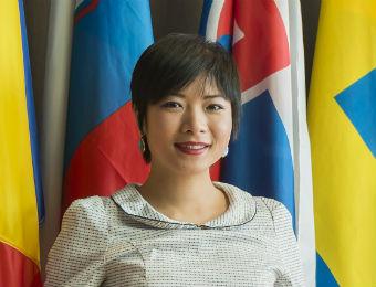 The image shows a woman smiling at the camera, with a background of various colourful national flags.